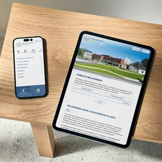 Smartphone und Tablet auf Holzbank zeigen responsive Website des Klinikverbunds Allgäu mit Startseite, Navigation und Standortwahl.