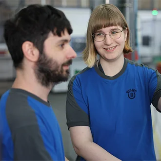Zwei junge Mitarbeitende der Dr. Werner Röhrs GmbH & Co. KG in blauer Arbeitskleidung bedienen gemeinsam ein modernes Bedienpanel in einer industriellen Produktionshalle – Fokus auf Teamarbeit, Digitalisierung und technisches Know-how in der Fertigung.