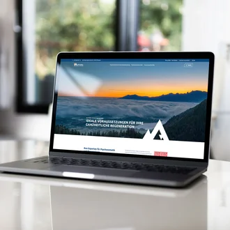 Laptop auf weißem Glastisch zeigt Startseite der Alpcura Fachklinik Allgäu mit Landschaftsmotiv und Slogan zur ganzheitlichen Regeneration.