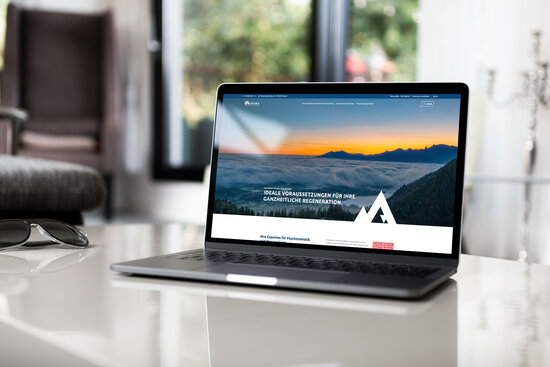 Laptop auf weißem Glastisch zeigt Startseite der Alpcura Fachklinik Allgäu mit Landschaftsmotiv und Slogan zur ganzheitlichen Regeneration.