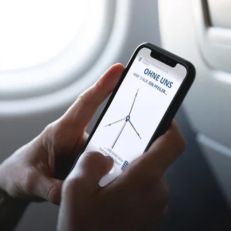 Person hält im Flugzeug ein Smartphone mit Anzeige einer Werbegrafik der Dr. Werner Röhrs GmbH & Co. KG mit einem Windrad und dem Text „Ohne uns wär’s nur ein Pfeiler…“ – Fokus auf Employer Branding, technische Bedeutung von Federn und Mobilität.