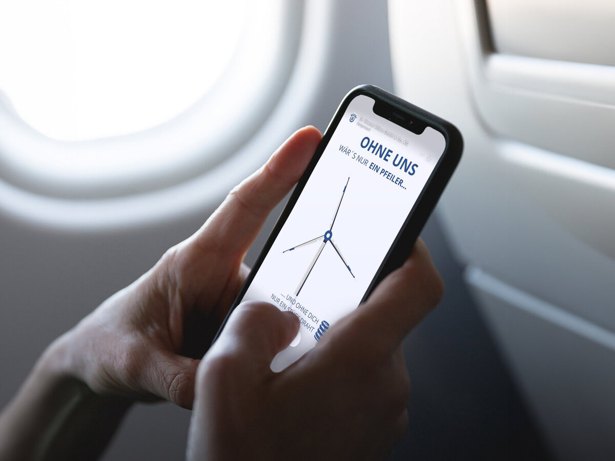 Person hält im Flugzeug ein Smartphone mit Anzeige einer Werbegrafik der Dr. Werner Röhrs GmbH & Co. KG mit einem Windrad und dem Text „Ohne uns wär’s nur ein Pfeiler…“ – Fokus auf Employer Branding, technische Bedeutung von Federn und Mobilität.