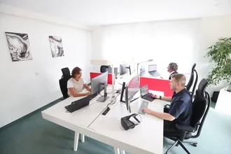 Denkströme Team arbeitet im hellen Großraumbüro an mehreren Monitoren und Laptops, getrennt durch rote Sichtschutzwände – Arbeitsplatzsituation für Website-Relaunch und digitale Projekte.