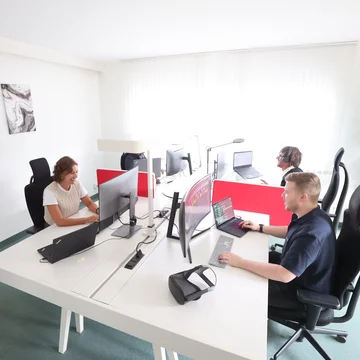 Denkströme Team arbeitet im hellen Großraumbüro an mehreren Monitoren und Laptops, getrennt durch rote Sichtschutzwände – Arbeitsplatzsituation für Website-Relaunch und digitale Projekte.