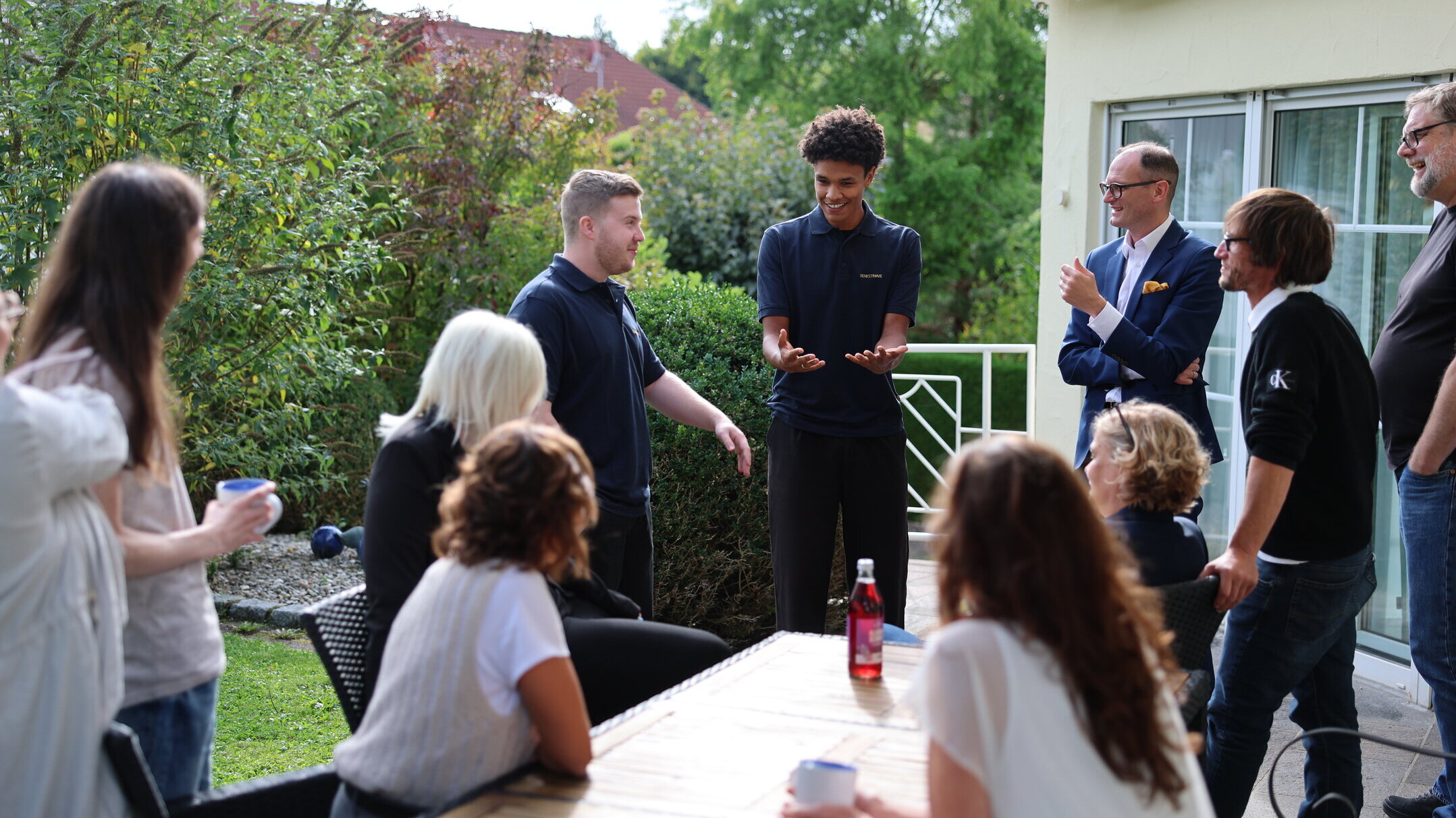 Team von Denkströme auf der Terrasse in Kempten: Mitarbeitende stehen und sitzen um einen Tisch, tauschen sich aus und trinken Kaffee im Gartenbereich.