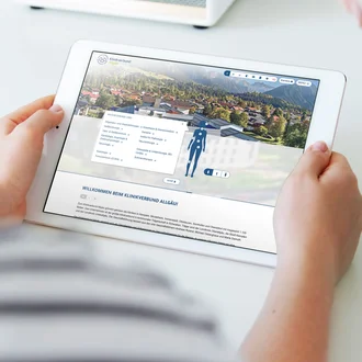 Person hält Tablet mit geöffneter Website des Klinikverbunds Allgäu, sichtbar ist interaktiver Körperkompass zur Fachbereichsauswahl und Standortansicht.