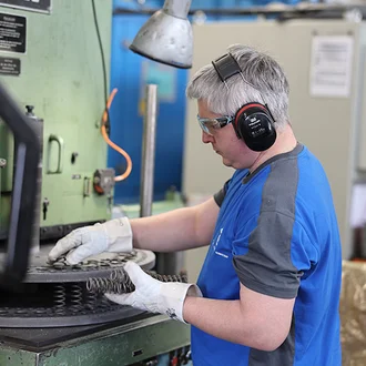 Mitarbeiter in der Produktion der Dr. Werner Röhrs GmbH & Co. KG arbeitet konzentriert an einer Industriemaschine mit Schutzbrille, Gehörschutz und Handschuhen – Fertigung technischer Federn unter Einhaltung hoher Sicherheits- und Qualitätsstandards.
