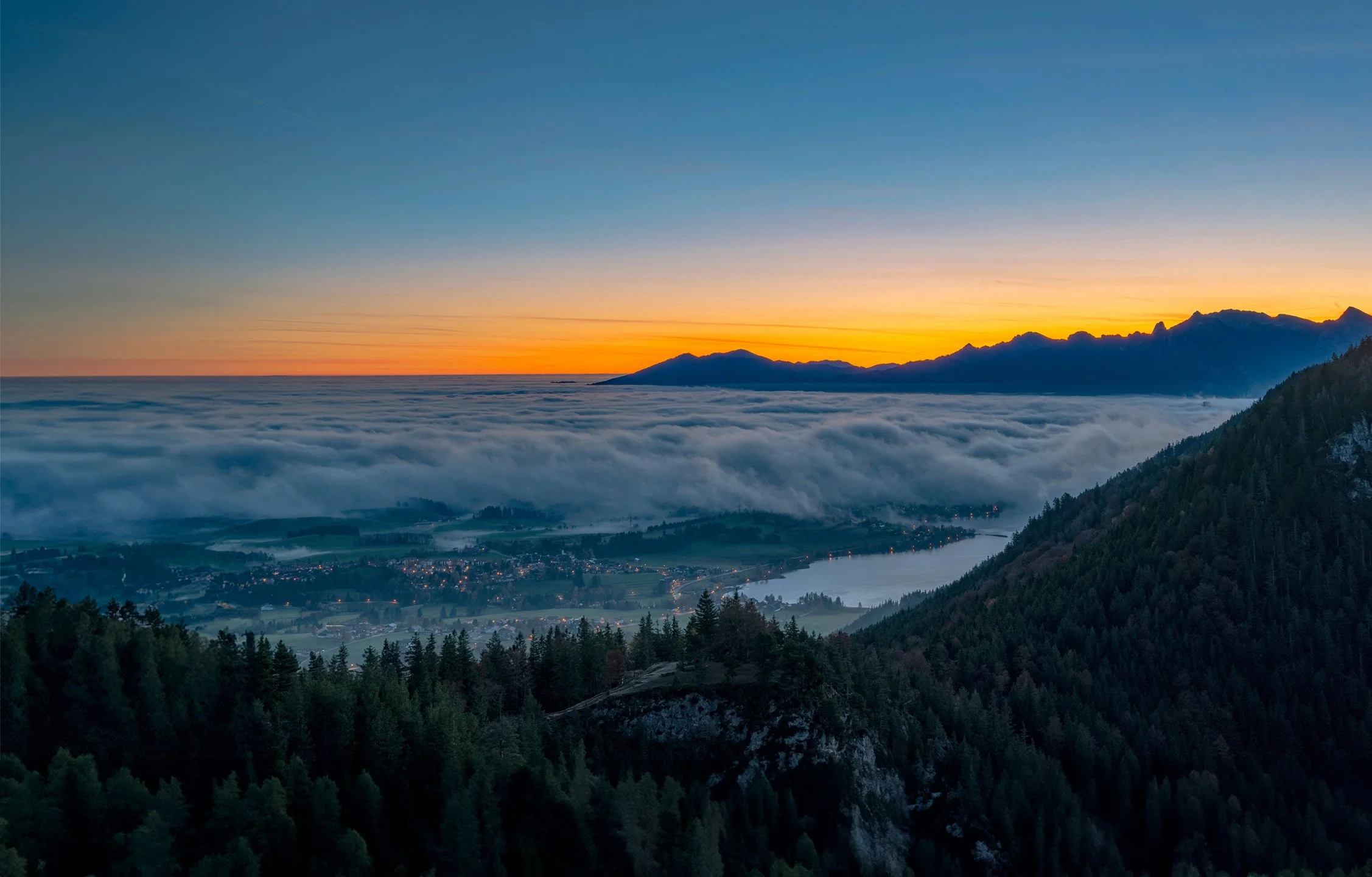 Nebelverhülltes Alpenvorland bei Sonnenaufgang mit Blick auf bewaldete Berghänge, See und beleuchtete Ortschaft im Tal.