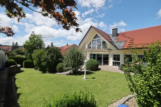 Außenansicht der Denkströme in Kempten von der Gartenseite: helles Bürogebäude mit rotem Ziegeldach, Terrasse, gepflegtem Rasen und Büschen unter blauem Himmel.
