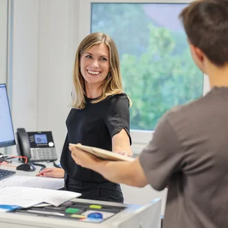 Lächelnde Mitarbeiterin am Büroarbeitsplatz übergibt Dokumente an Kollegen – Teamarbeit im kaufmännischen Bereich, moderne Büroausstattung mit Bildschirmarbeitsplatz