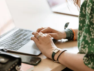 Teilansicht eines Laptops auf einem Schreibtisch, aus dem leuchtende digitale Symbole und Interface-Elemente wie Suchleisten, Herzen, Chatblasen, Kamera-, Cloud- und Nutzer-Icons holografisch hervorgehoben sind