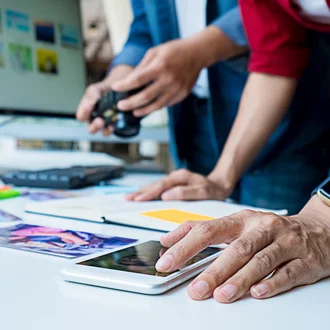 Zwei Personen arbeiten an einem Kreativprojekt mit Kamera, Smartphone, Farbkarten und Fotos am Schreibtisch – im Hintergrund ein großer Bildschirm mit Bildauswahl