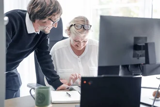 Zwei Mitarbeitende bei Denkströme arbeiten gemeinsam am Schreibtisch: Eine Person steht daneben und zeigt auf den Bildschirm, die andere sitzt am Computer und bedient die Maus.