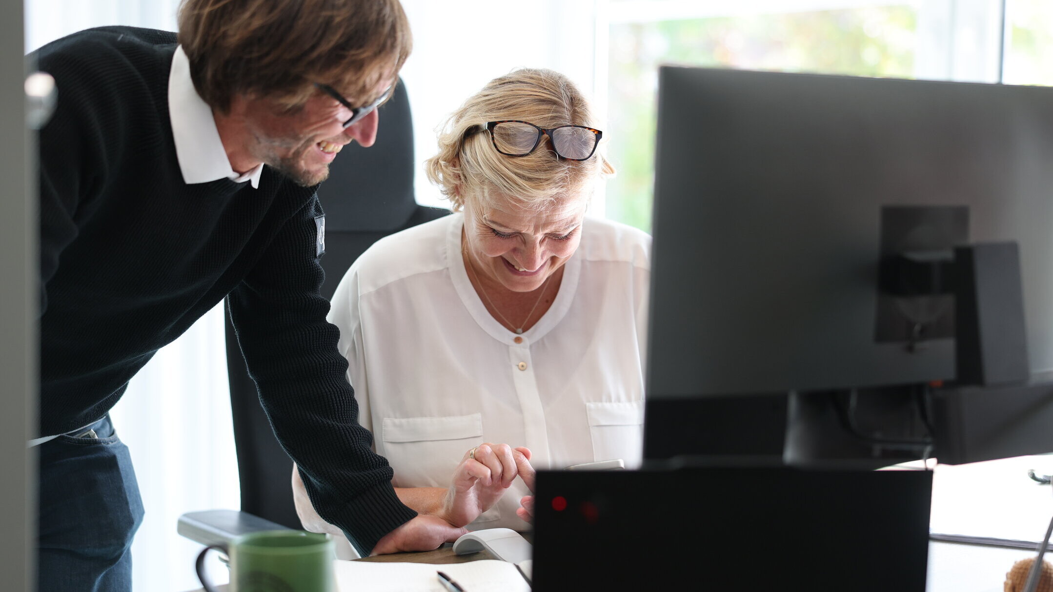 Zwei Mitarbeitende bei Denkströme arbeiten gemeinsam am Schreibtisch: Eine Person steht daneben und zeigt auf den Bildschirm, die andere sitzt am Computer und bedient die Maus.