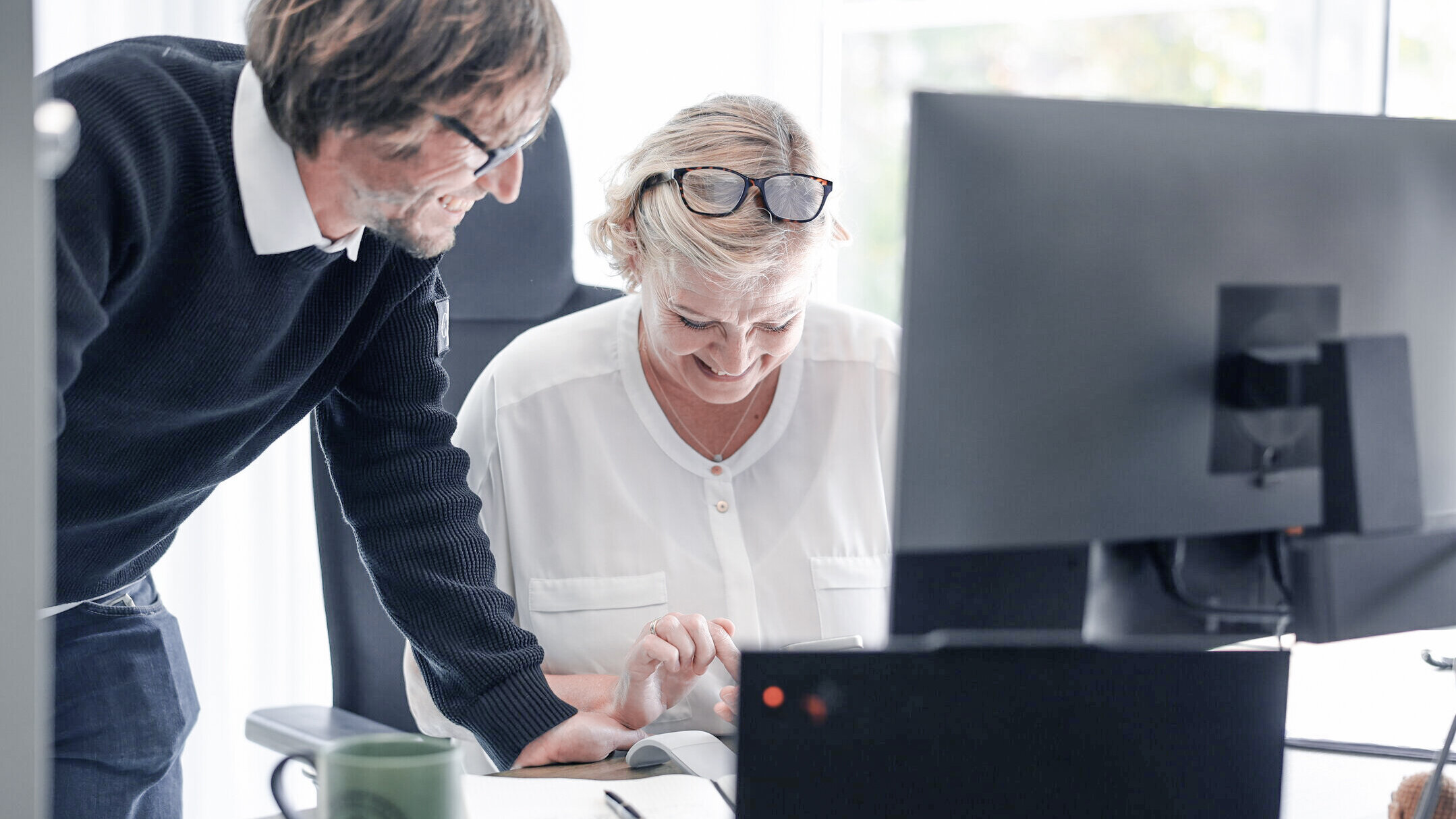 Zwei Mitarbeitende bei Denkströme arbeiten gemeinsam am Schreibtisch: Eine Person steht daneben und zeigt auf den Bildschirm, die andere sitzt am Computer und bedient die Maus.