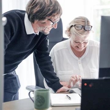 Zwei Mitarbeitende bei Denkströme arbeiten gemeinsam am Schreibtisch: Eine Person steht daneben und zeigt auf den Bildschirm, die andere sitzt am Computer und bedient die Maus.