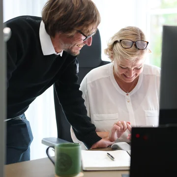 Zwei Mitarbeitende bei Denkströme arbeiten gemeinsam am Schreibtisch: Eine Person steht daneben und zeigt auf den Bildschirm, die andere sitzt am Computer und bedient die Maus.