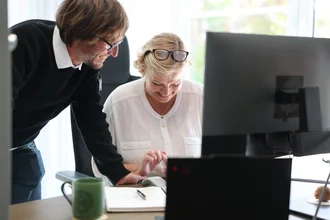Zwei Mitarbeitende bei Denkströme arbeiten gemeinsam am Schreibtisch: Eine Person steht daneben und zeigt auf den Bildschirm, die andere sitzt am Computer und bedient die Maus.