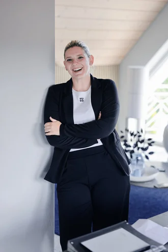 Teresa Schmid von Denkströme steht lächelnd im Büroflur, lehnt an einer Wand und hat die Arme verschränkt.