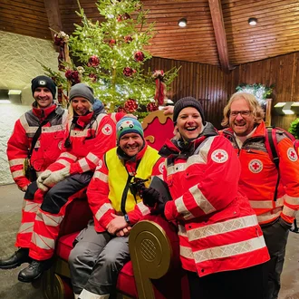 Gruppe fröhlicher BRK-Einsatzkräfte in roten Uniformen vor festlich geschmücktem Weihnachtsbaum mit Lichterketten und Kugeln – Gemeinschaft und Engagement im weihnachtlichen Einsatz