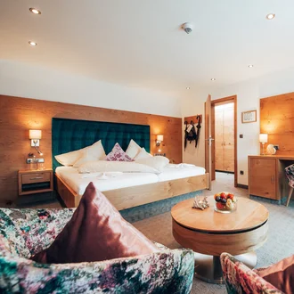 Komfortables Hotelzimmer mit Kingsize-Bett, großem TV, floralen Sesseln und Holzdesign im modernen Alpenstil