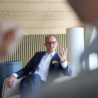 Beratungsgespräch bei Denkströme: Ein Manager im Anzug sitzt im Sessel, spricht lächelnd und gestikuliert; im Vordergrund ist unscharf ein Zuhörender zu sehen.