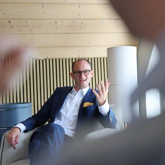 Beratungsgespräch bei Denkströme: Ein Manager im Anzug sitzt im Sessel, spricht lächelnd und gestikuliert; im Vordergrund ist unscharf ein Zuhörender zu sehen.