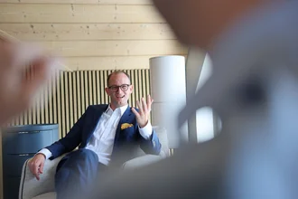 Beratungsgespräch bei Denkströme: Ein Manager im Anzug sitzt im Sessel, spricht lächelnd und gestikuliert; im Vordergrund ist unscharf ein Zuhörender zu sehen.