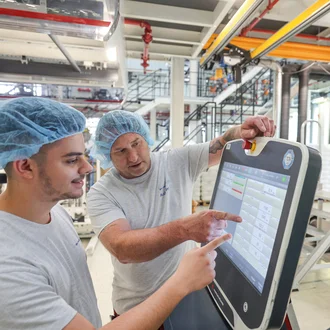 Zwei Mitarbeiter mit Haarnetz bedienen eine industrielle Maschinensteuerung über ein Touchpanel – moderne Produktionstechnik in der Kunststoffverarbeitung