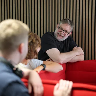 Denkströme Team im Gespräch in der roten Lounge im Büro; ein Mitarbeiter lehnt an der Sitzbank und diskutiert mit Kolleginnen und Kollegen zur Social-Media-Strategie.