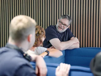 Denkströme Team im Gespräch in der roten Lounge im Büro; ein Mitarbeiter lehnt an der Sitzbank und diskutiert mit Kolleginnen und Kollegen zur Social-Media-Strategie.
