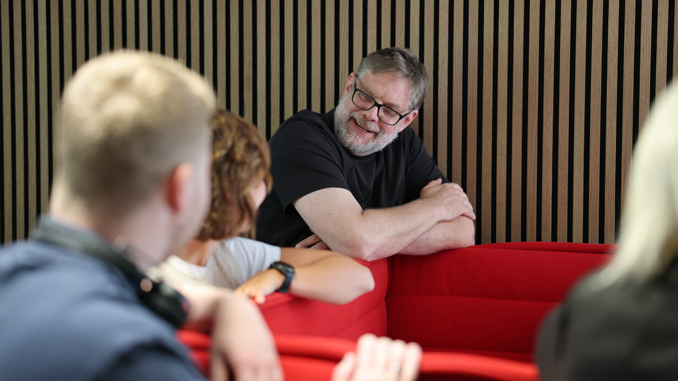 Denkströme Team im Gespräch in der roten Lounge im Büro; ein Mitarbeiter lehnt an der Sitzbank und diskutiert mit Kolleginnen und Kollegen zur Social-Media-Strategie.