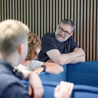 Denkströme Team im Gespräch in der roten Lounge im Büro; ein Mitarbeiter lehnt an der Sitzbank und diskutiert mit Kolleginnen und Kollegen zur Social-Media-Strategie.