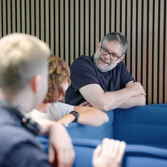 Denkströme Team im Gespräch in der roten Lounge im Büro; ein Mitarbeiter lehnt an der Sitzbank und diskutiert mit Kolleginnen und Kollegen zur Social-Media-Strategie.