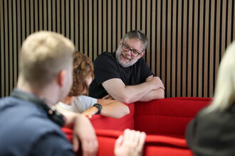 Denkströme Team im Gespräch in der roten Lounge im Büro; ein Mitarbeiter lehnt an der Sitzbank und diskutiert mit Kolleginnen und Kollegen zur Social-Media-Strategie.