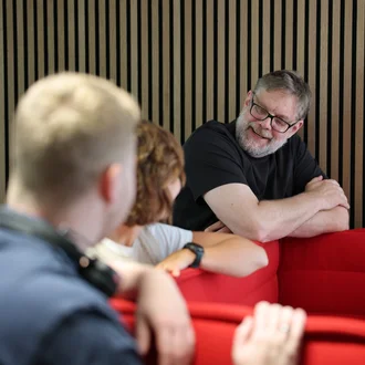 Denkströme Team im Gespräch in der roten Lounge im Büro; ein Mitarbeiter lehnt an der Sitzbank und diskutiert mit Kolleginnen und Kollegen zur Social-Media-Strategie.