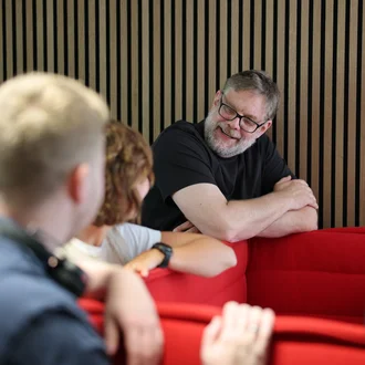Denkströme Team im Gespräch in der roten Lounge im Büro; ein Mitarbeiter lehnt an der Sitzbank und diskutiert mit Kolleginnen und Kollegen zur Social-Media-Strategie.