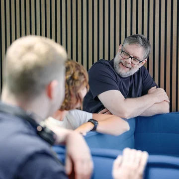 Denkströme Team im Gespräch in der roten Lounge im Büro; ein Mitarbeiter lehnt an der Sitzbank und diskutiert mit Kolleginnen und Kollegen zur Social-Media-Strategie.