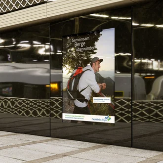 Junger Auszubildender mit Rucksack blickt optimistisch in die Allgäuer Berglandschaft – Plakatmotiv der Arbeitgeberkampagne „Gemeinsam Berge überwinden“ der Fachklinik Allgäu in einem modernen Bahnhofsumfeld.