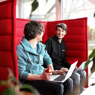 Zwei Denkströme Mitarbeiter im Gespräch in modernem Büro mit roten Akustik-Sesseln, arbeiten gemeinsam mit Laptops, kreativer Austausch