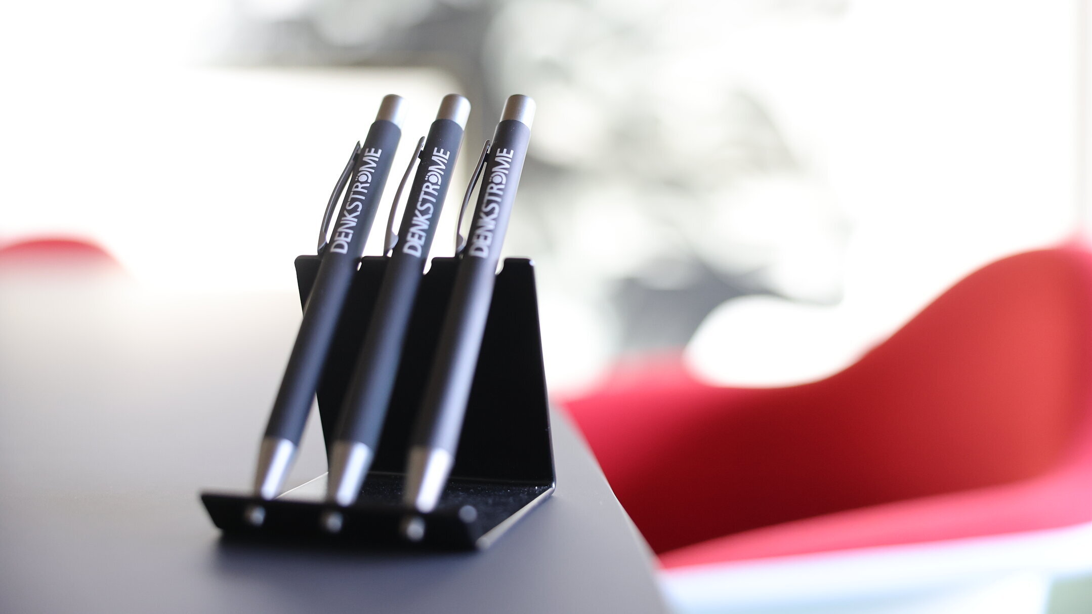 Drei Denkströme-Kugelschreiber stehen in einem Stifthalter auf einem Konferenztisch; im Hintergrund ist ein roter Stuhl unscharf zu sehen.