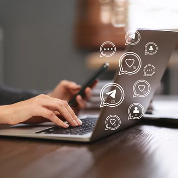 Person tippt auf einem Laptop und hält ein Smartphone, umgeben von digitalen Symbolen für Likes, Nachrichten und Follower