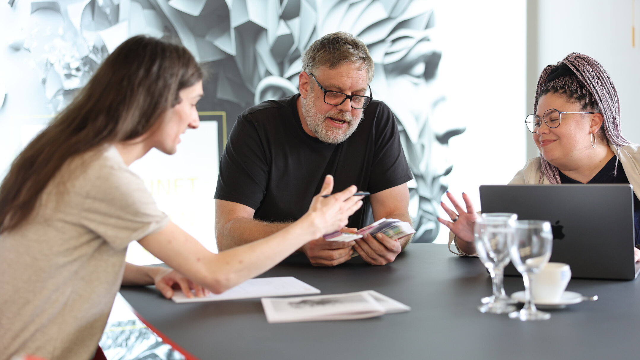 Drei Mitarbeitende aus der Grafikabteilung von Denkströme besprechen Corporate-Design-Entwürfe am Konferenztisch; eine Person arbeitet am Laptop, zwei zeigen auf Vorlagen.