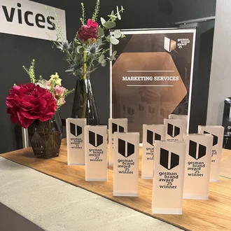 Ausgezeichnete Markenarbeit: Mehrere Trophäen des German Brand Award 2025 in der Kategorie „Marketing Services“ stehen auf einem modernen Sideboard neben stilvoll arrangierten Blumenvasen. Die Szene zeigt die hochwertige Inszenierung der Preisverleihung, bei der exzellente Markenführung gewürdigt wird.