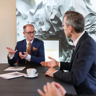 Beratungsgespräch bei Denkströme: Drei Personen diskutieren am Konferenztisch; ein Manager im blauen Anzug gestikuliert, auf dem Tisch stehen Kaffeetasse und Unterlagen.