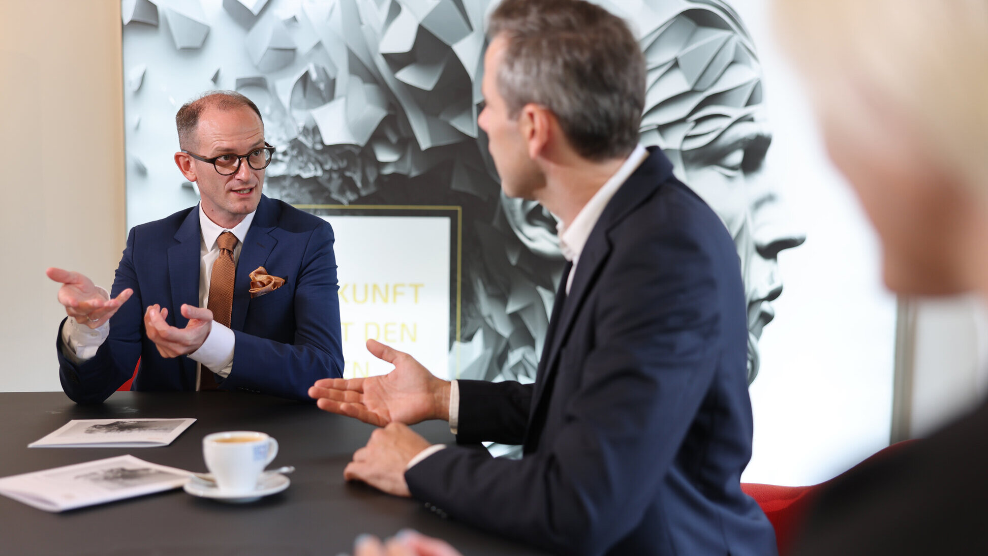 Beratungsgespräch bei Denkströme: Drei Personen diskutieren am Konferenztisch; ein Manager im blauen Anzug gestikuliert, auf dem Tisch stehen Kaffeetasse und Unterlagen.