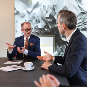 Beratungsgespräch bei Denkströme: Drei Personen diskutieren am Konferenztisch; ein Manager im blauen Anzug gestikuliert, auf dem Tisch stehen Kaffeetasse und Unterlagen.
