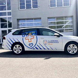 Weißer Dienstwagen mit Logo des Allgemeinen Krankenhauses Celle vor moderner Gebäudefassade mit Glas und Metallpanelen