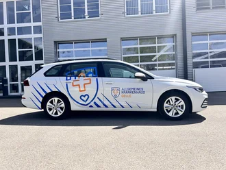 Weißer Dienstwagen mit Logo des Allgemeinen Krankenhauses Celle vor moderner Gebäudefassade mit Glas und Metallpanelen