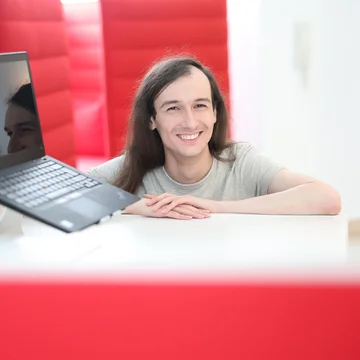 Mitarbeiter aus der Grafikabteilung von Denkströme lächelt im Büro hinter einem Schreibtisch; Laptop und Monitor stehen im Vordergrund, rote Akustikelemente im Hintergrund.